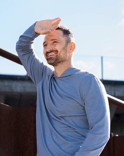 A smiling man stands outdoors on a sunny day, shielding his eyes from the sun with one hand. He is wearing a long-sleeve, dusty blue hoodie. In the background, there is a rusty metal structure and part of an elevated concrete overpass.
