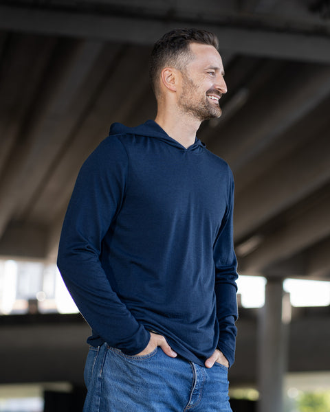 A smiling man stands outdoors with both hands in the front pockets of his blue jeans. He is wearing a long-sleeve, navy blue hoodie. The background shows the underside of a large concrete overpass structure.