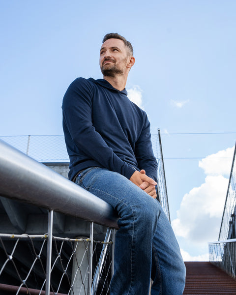 A man with a short beard sits casually on the thick metal railing of an outdoor walkway or bridge, looking off to the left. He is wearing a long-sleeve navy blue hoodie and blue jeans. The background features a bright blue sky with white clouds and the modern architectural elements of the walkway.