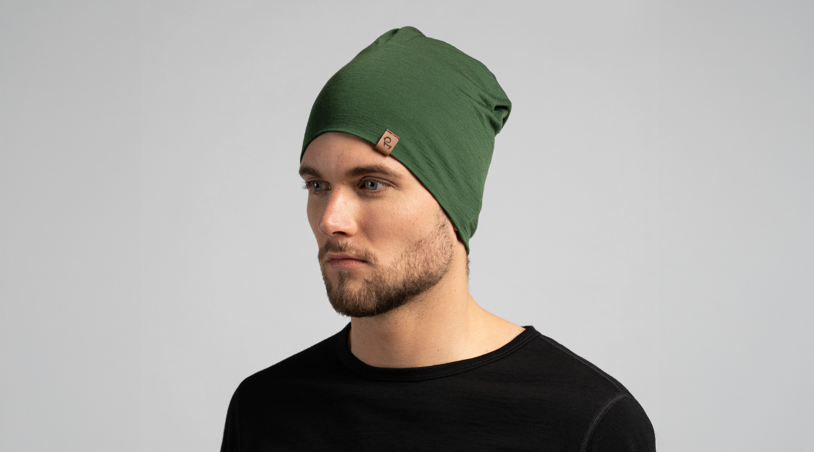 A close-up portrait shows a man wearing a snug green beanie featuring a small square leather logo tag. He is looking slightly off-camera while wearing a simple black crew-neck top against a light background.
