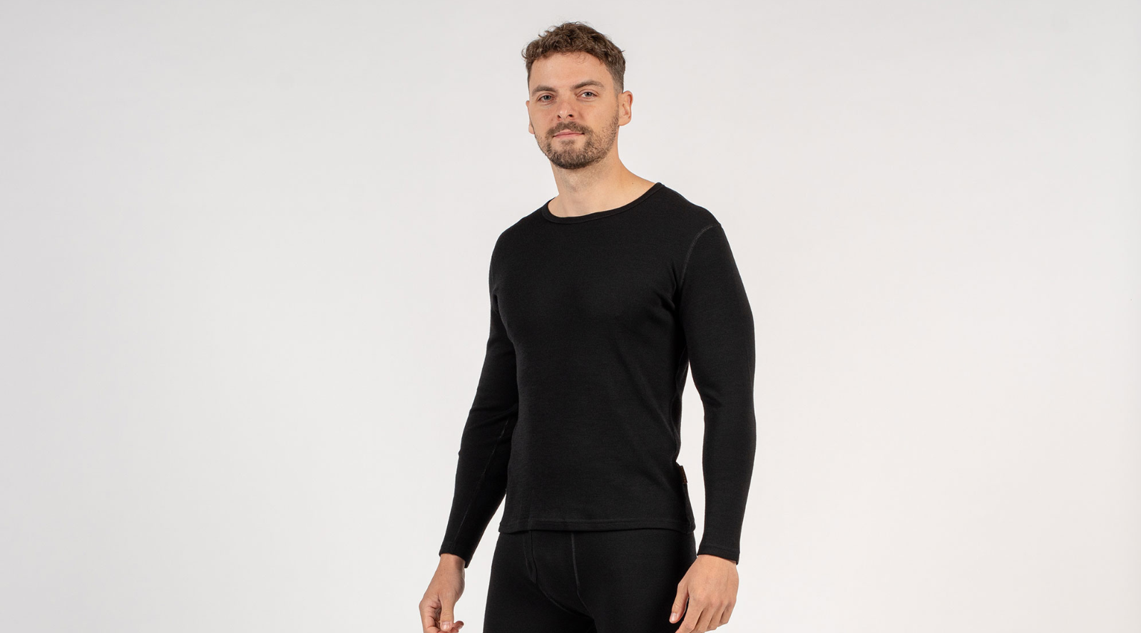 A man with a short beard stands facing forward in a black long-sleeve mid-layer top and black thermal leggings. The image is a clear studio shot set against a clean, light gray background.