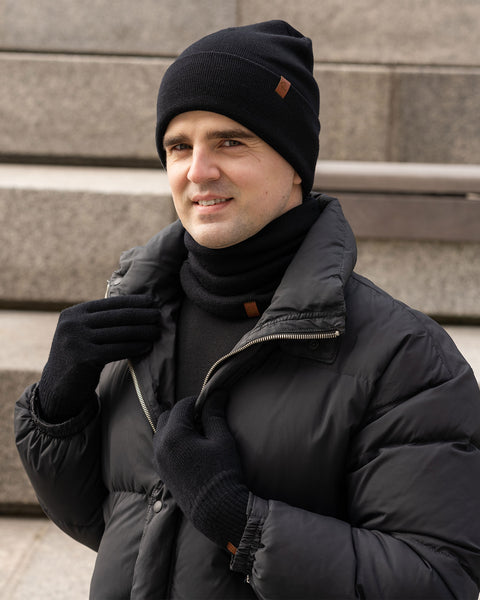 A person with light skin and dark hair is smiling and looking towards the camera. They are wearing a black knit beanie with a small brown tag, a black knit neck gaiter with a similar tag, black knit gloves, and a black puffer jacket. They are standing outdoors on what appears to be concrete steps.