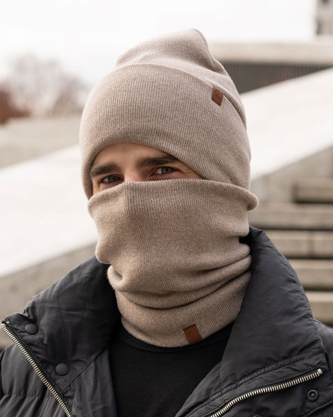 A person with light brown hair and eyes is shown outdoors, wearing a creamy beige knit beanie pulled down to their eyebrows and a matching neck gaiter covering their mouth and nose. They are wearing a black puffer jacket. The beanie has a small brown tag on the side. The background is blurred, suggesting an urban environment with steps or concrete structures. Only the person's eyes are visible above the neck gaiter.