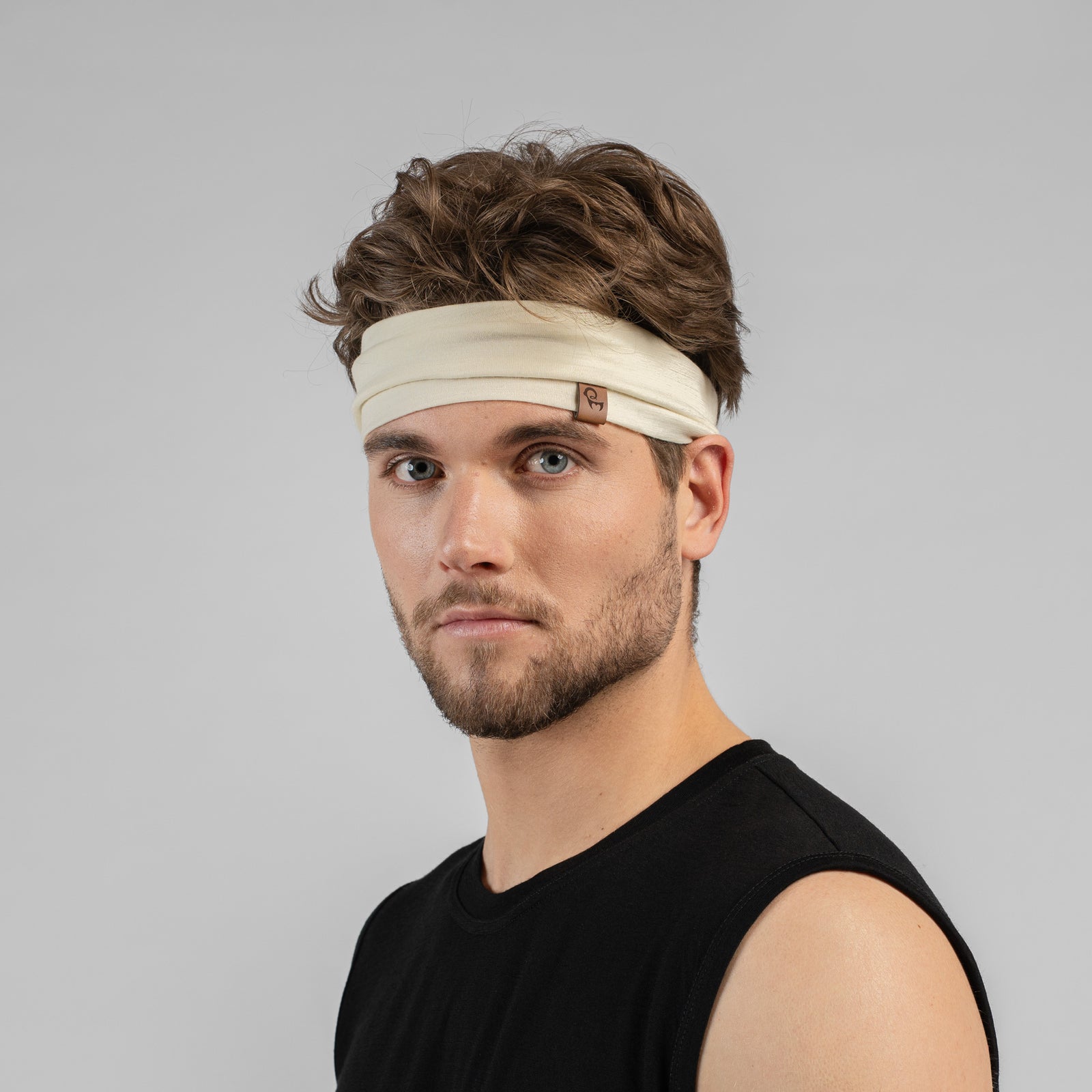 An isolated product shot of a wide, natural off-white fabric headband against a plain white background. The headband features a small, square brown patch with a stylized logo stitched onto the center front. The material appears soft with a subtle, fine-knit texture.