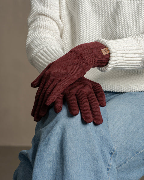 Hands wearing deep cherry red merino wool knit gloves, styled with a cream sweater, highlighting warmth and refined knit texture.