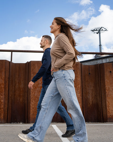 A man and a woman walk side-by-side outdoors on a paved surface. The woman is in the foreground wearing a beige hoodie and light blue jeans, with her hair blowing in the wind. The man is slightly behind her, wearing a navy blue hoodie and blue jeans. They are walking past a rusty corrugated metal wall.