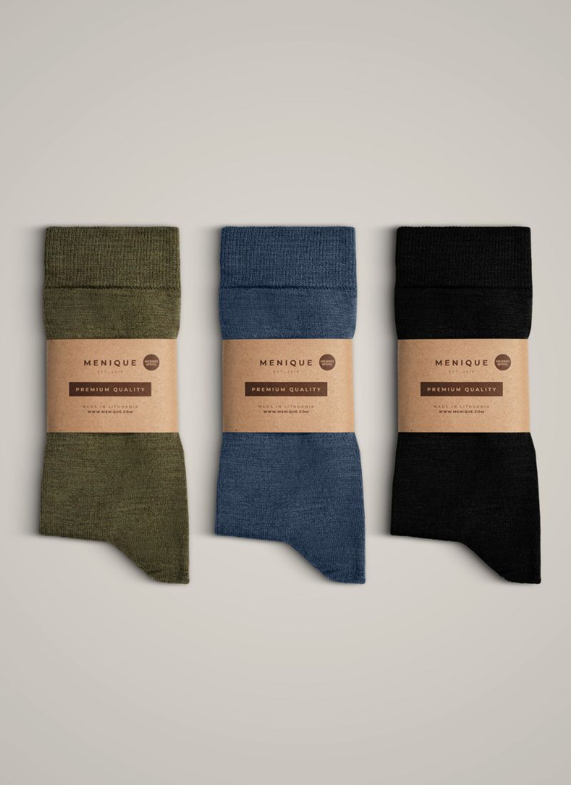Three pairs of folded knit socks in olive green, slate blue, and black are arranged vertically against a neutral gray background. Each pair is wrapped in a brown kraft paper sleeve featuring the "MENIQUE" brand logo and "PREMIUM QUALITY" text.