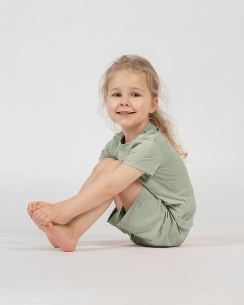 A studio portrait of a smiling young blonde girl sitting barefoot on the floor. She is angled slightly to the left, looking directly at the camera with her knees pulled up towards her chest and her hands holding her toes. She is wearing a matching set of a light sage green short-sleeved t-shirt and shorts against a plain light grey background.