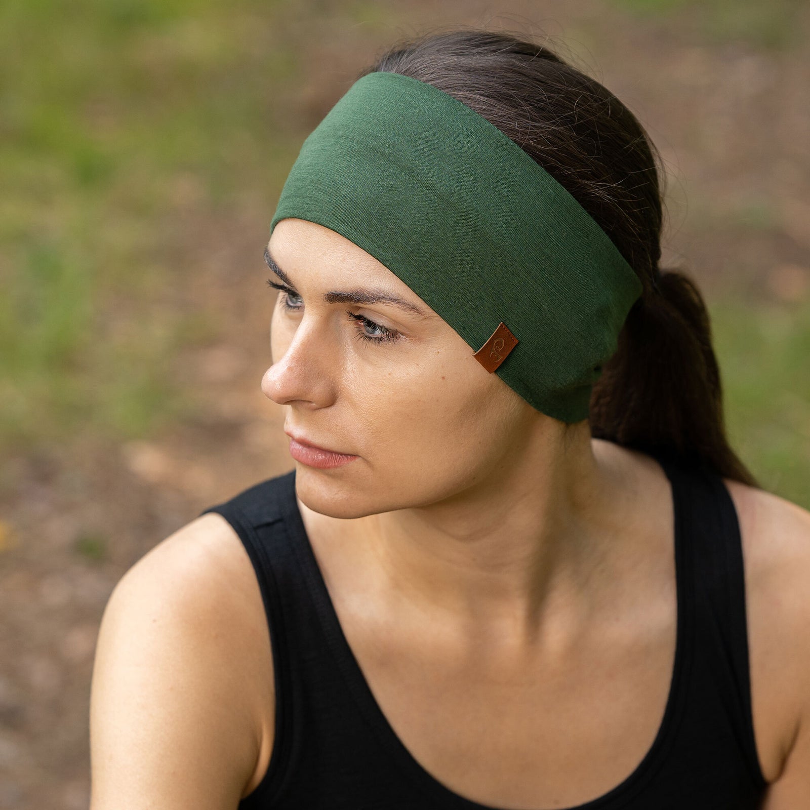 The picture features a couple of people - man and woman - doing leg warm-up exercises. They are stretching in a forest, preparing for a run. They are wearing sleeveless black merino two-piece sets and dark green unisex merino headbands.