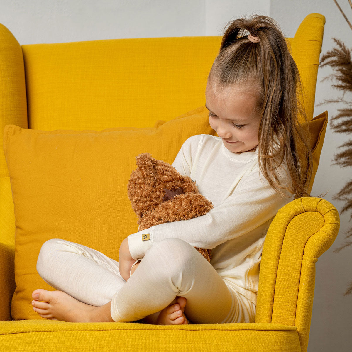 A girl sitting in a yellow armchair, with her legs crossed, wearing the natural color menique long sleeve merino 160gsm top in natural color. 