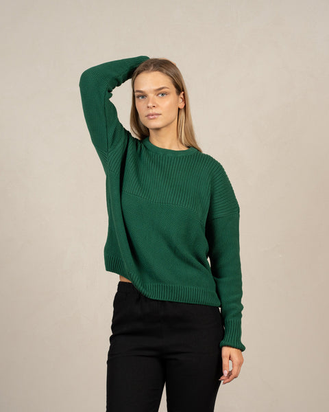 A young blond woman wearing the menique combed cotton sweater SONATA in green color, with one of her hands behind her head.