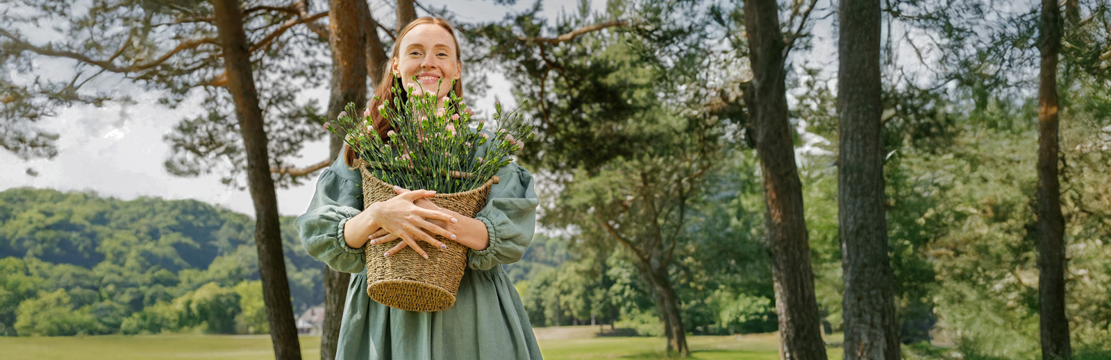 Woman holding basket of flowers in nature, wearing linen dress in summer landscape