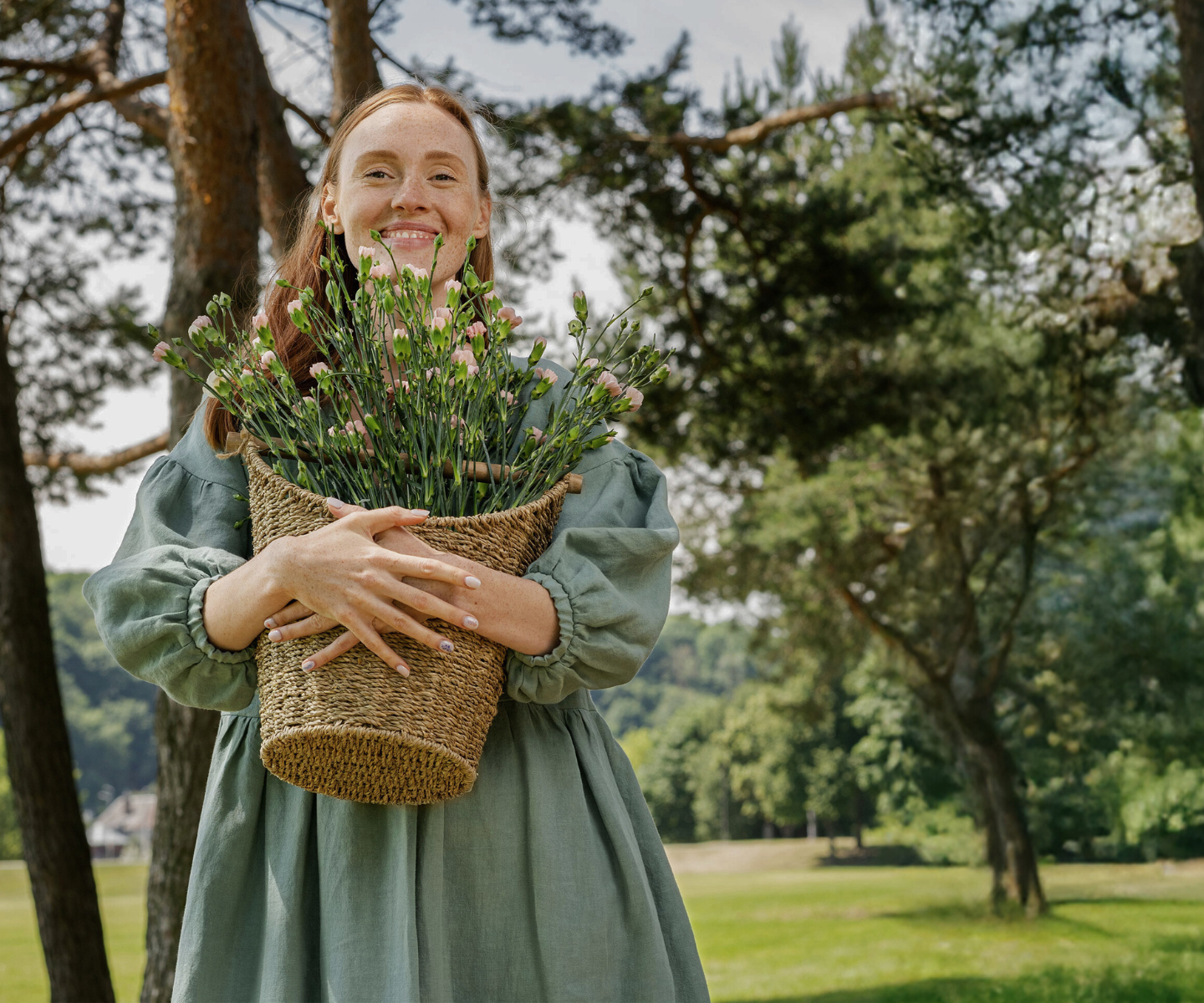 Woman holding basket of flowers in nature, wearing linen dress in summer landscape