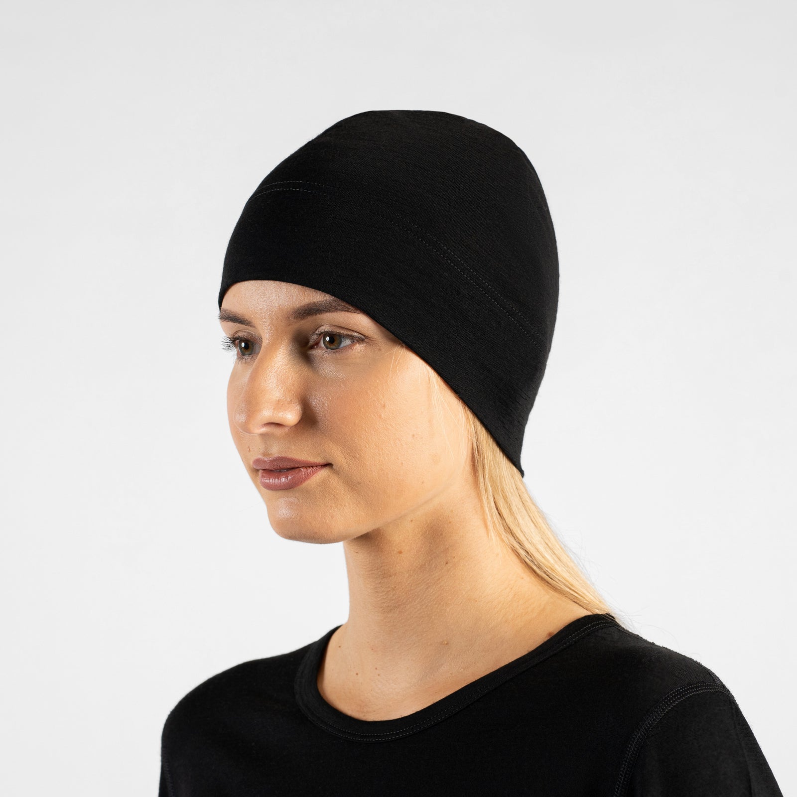 A studio portrait of a woman in a profile view, looking off to the left side of the frame. She is wearing a fitted, dark red (royal cherry) skull cap over her blonde hair and a black top. The background is a solid light grey.