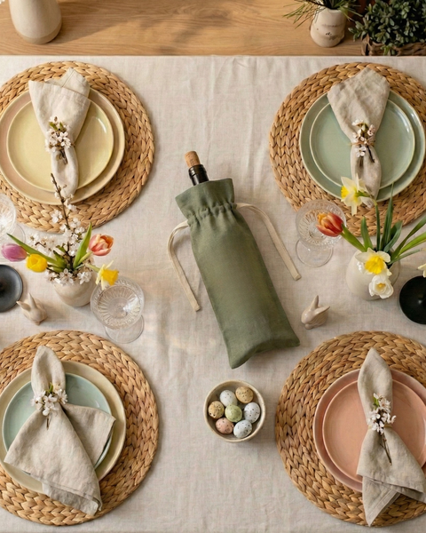 Overhead view of a spring table setting with woven placemats, pastel plates, flowers, and a wine bottle in a green linen drawstring bag.