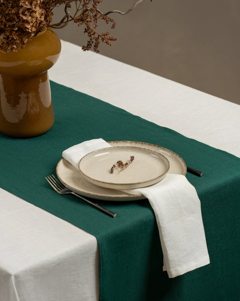 A tabletop covered with a linen tablecloth, with a vase and a single plate set on top of it. By the sides of the plates there are cutlery items, and there is a linen napkin in pure white color on top.