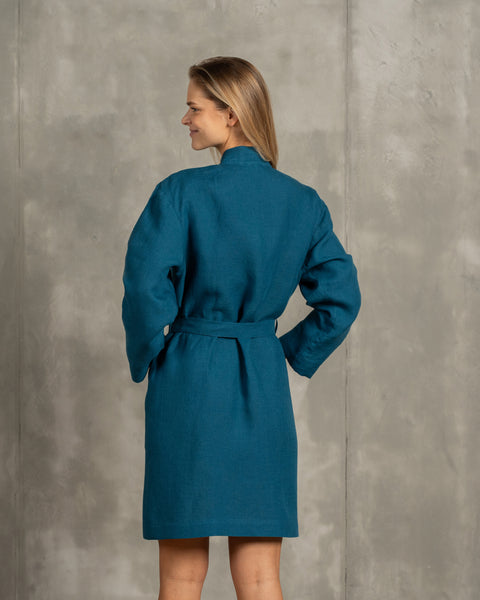 The menique women's linen bathrobe in cobalt blue color. The woman is standing in front of a gray wall and showing the bathrobe from the back. The adjustable tie is visible from the back.