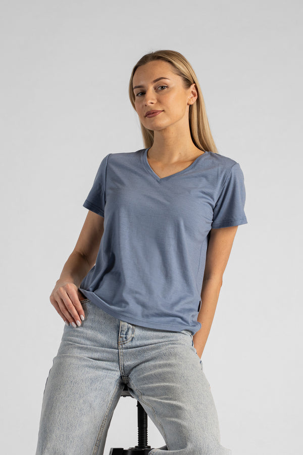 A studio portrait of a blonde woman sitting on a stool and looking directly at the camera. She is leaning back slightly, with her hands resting on her lap. She is wearing a regular fit, dusty blue short-sleeved v-neck t-shirt and light-wash blue jeans against a plain light grey background.
