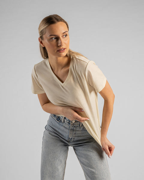 A studio portrait of a blonde woman leaning slightly forward and looking off to the right, gently pulling at the bottom hem of her shirt. She is wearing a natural off-white short-sleeved V-neck t-shirt and light wash denim jeans against a plain light grey background.