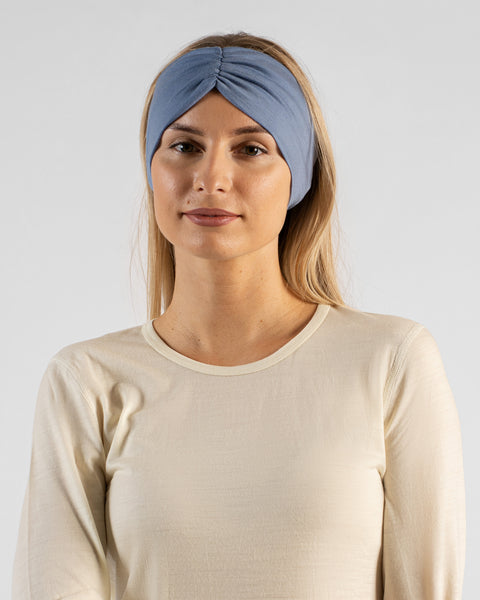 A close-up studio portrait of a blonde woman looking directly at the camera. She is wearing a light beige long-sleeved shirt and a wide dusty blue headband with a gathered twist detail at the front, against a plain light grey background.