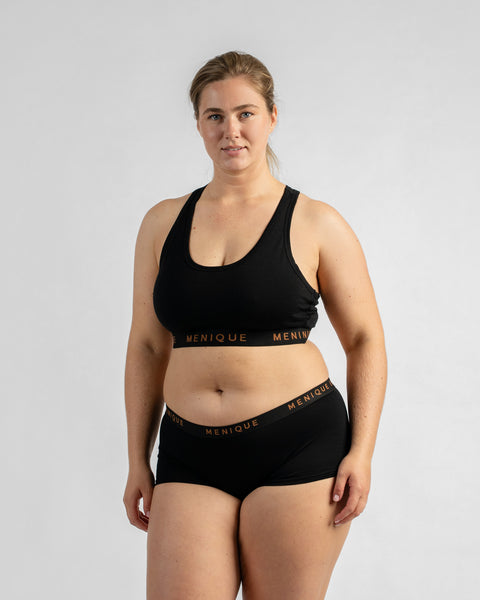 Front view of a model standing with her arms at her sides, wearing a black sports bra and matching boy-short underwear, both featuring a black elastic band.
