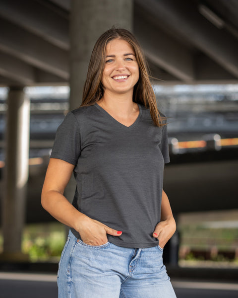 A close-up variation of a woman standing under a concrete structure, smiling warmly directly at the camera. She is wearing a charcoal grey v-neck t-shirt and light blue jeans, with her hands tucked into her pockets.