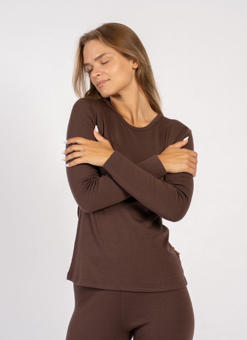 A woman with her eyes closed comfortably hugs herself while wearing a brown long-sleeve top and matching bottoms. The studio shot is set against a simple light gray backdrop to highlight the fit of the garments.