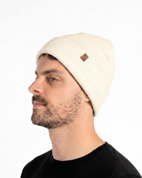 A profile portrait of a bearded man wearing a cream-colored ribbed knit beanie hat and a black shirt.