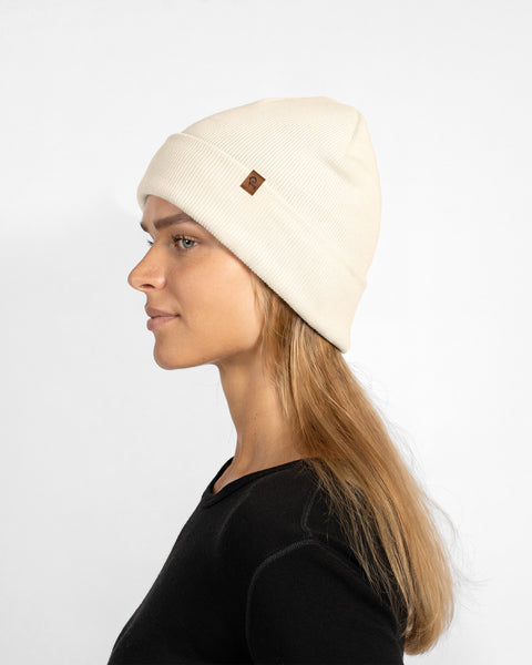 A profile portrait of a blonde woman wearing a cream-colored ribbed knit beanie hat and a black long-sleeved top.
