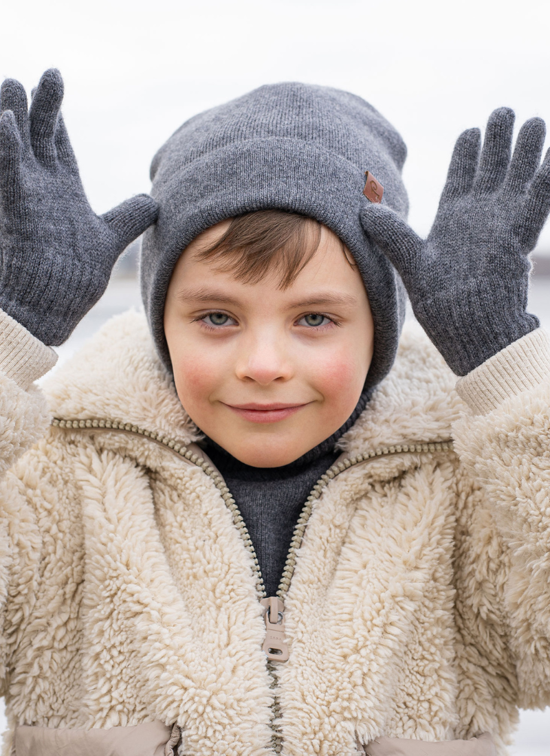 A young boy smiles playfully while wearing a grey knit beanie, matching gloves, and a textured cream-colored sherpa jacket. He holds his gloved hands up beside his head against a soft, out-of-focus outdoor background.