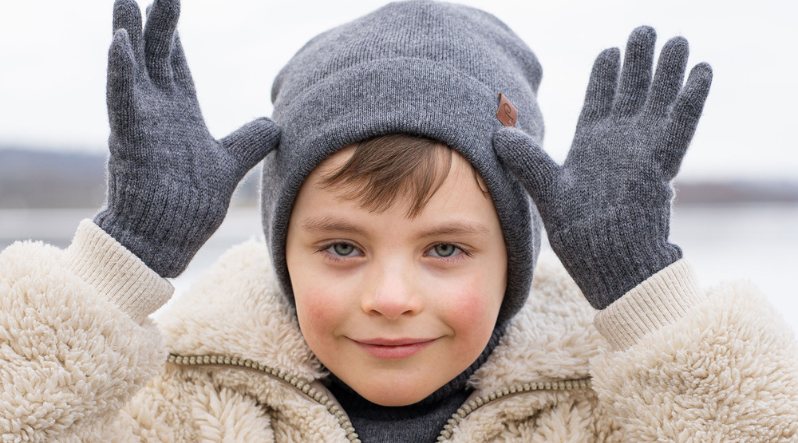 A young boy smiles playfully while wearing a grey knit beanie, matching gloves, and a textured cream-colored sherpa jacket. He holds his gloved hands up beside his head against a soft, out-of-focus outdoor background.