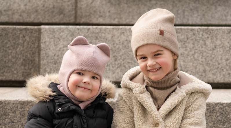 Two young children sit outdoors against a backdrop of light gray stone steps, bundled up in warm winter clothing. The child on the left is wearing a black puffy coat with a faux-fur hood and a dusty pink knit balaclava that features small animal ears on top, while the child on the right smiles brightly wearing a cream-colored textured sherpa coat, a beige ribbed turtleneck, and a matching beige beanie.