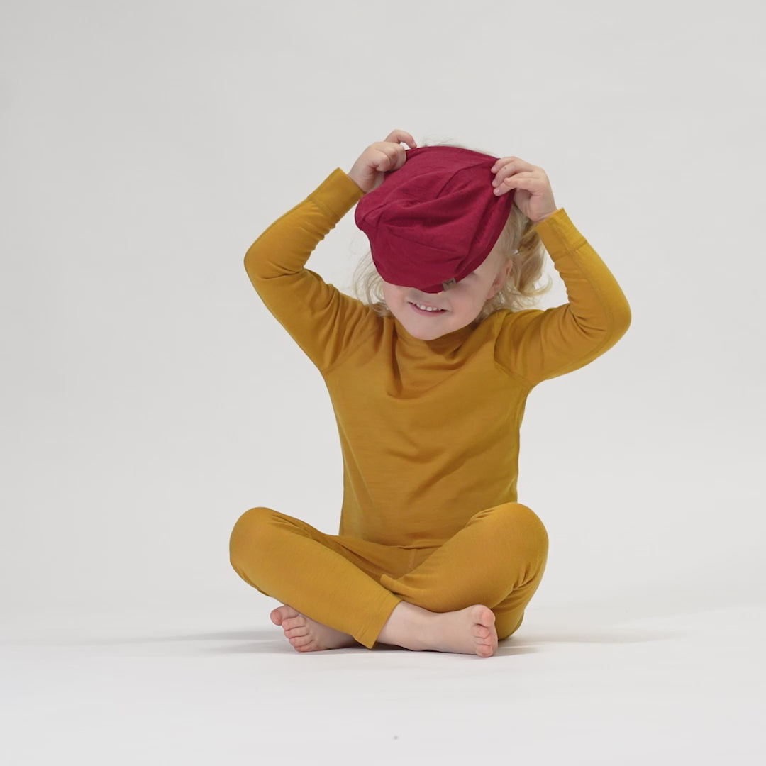 A smiling child with light, curly hair wears a royal cherry beanie and a yellow long-sleeved shirt. The child is resting their chin on their clasped hands and looking up at the camera. The background is white. 
