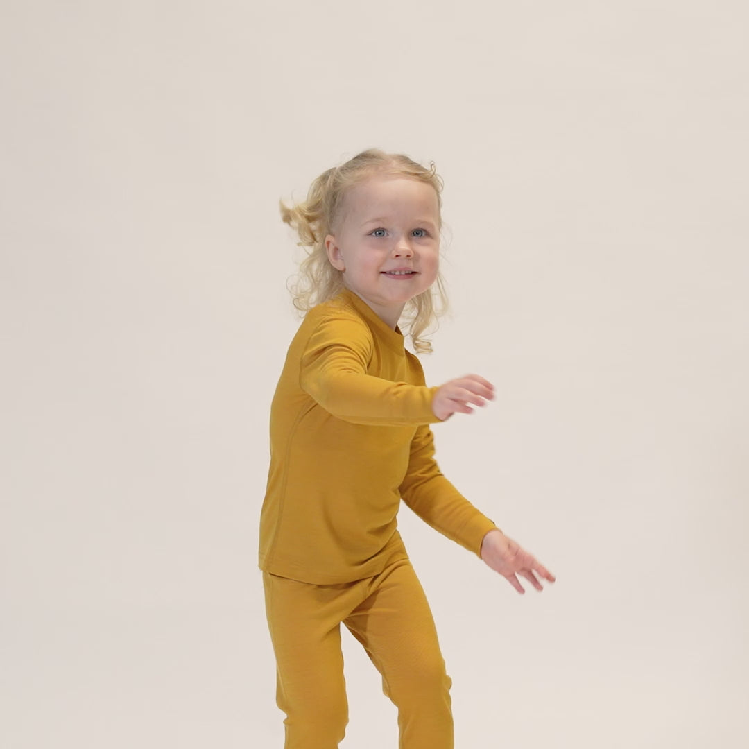 A child with curly blonde hair styled in pigtails, wearing a yellow long-sleeved top and matching pants, is captured mid-movement against a white background. 