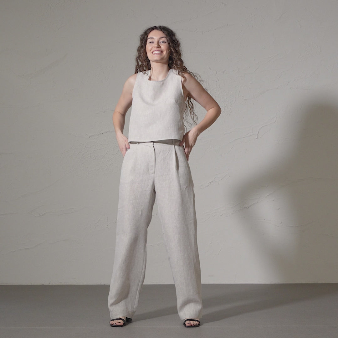 A smiling woman with long, curly brown hair models a linen sleeveless top and matching menique linen wide-legged pants, posing with her hands on her hips in a short video against a textured off-white wall.