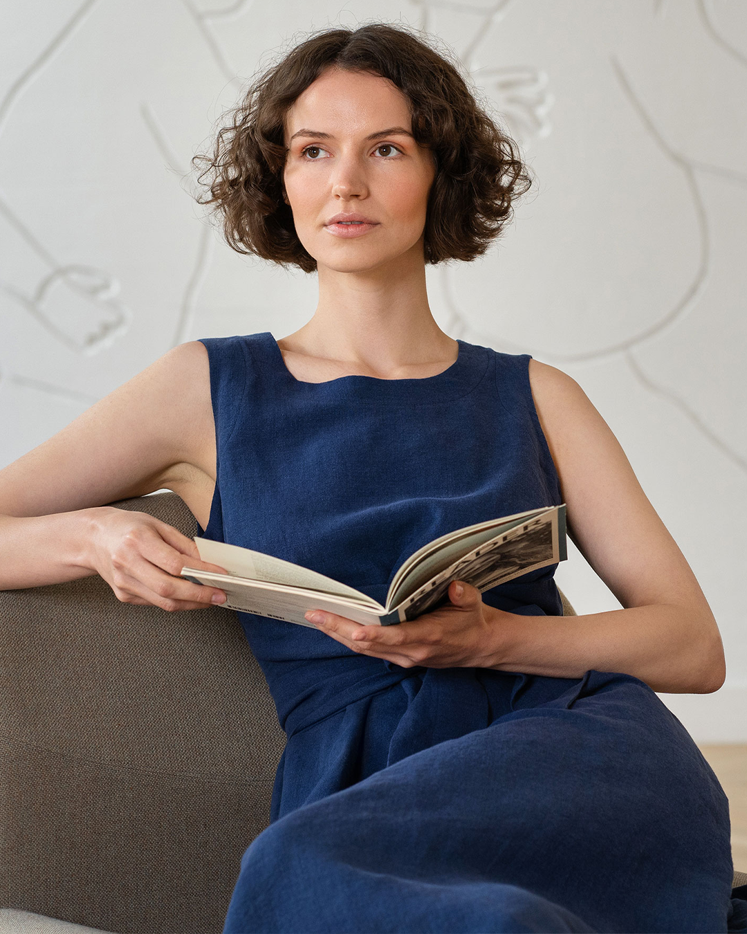 A woman with short dark hair sits on a brown sofa wearing a sleeveless navy blue dress while holding an open book.