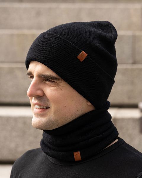 A young man, smiling faintly, is viewed in profile, looking to the right of the frame. He is wearing a black knitted beanie and a matching black gaiter. Both items have a small, rectangular brown leather patch with a Menique logo visible. He has short dark hair and appears to be outdoors, with a blurred background of stone steps or a building.