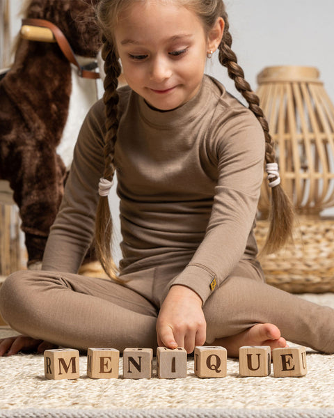 a girl wearing the menique Kids' Merino 160 Long Sleeve Crew in Beige color. She is playing with wooden  letter cubes on the carpet.